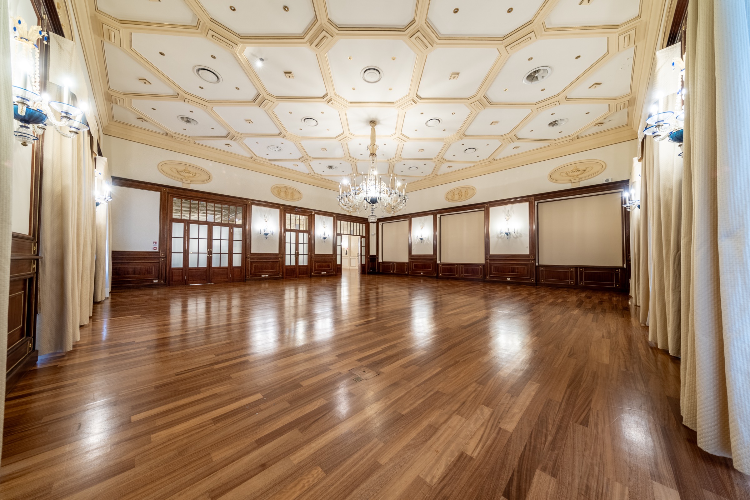Three ornate spherical chandeliers hanging in a grand stone-arched hallway with large windows.